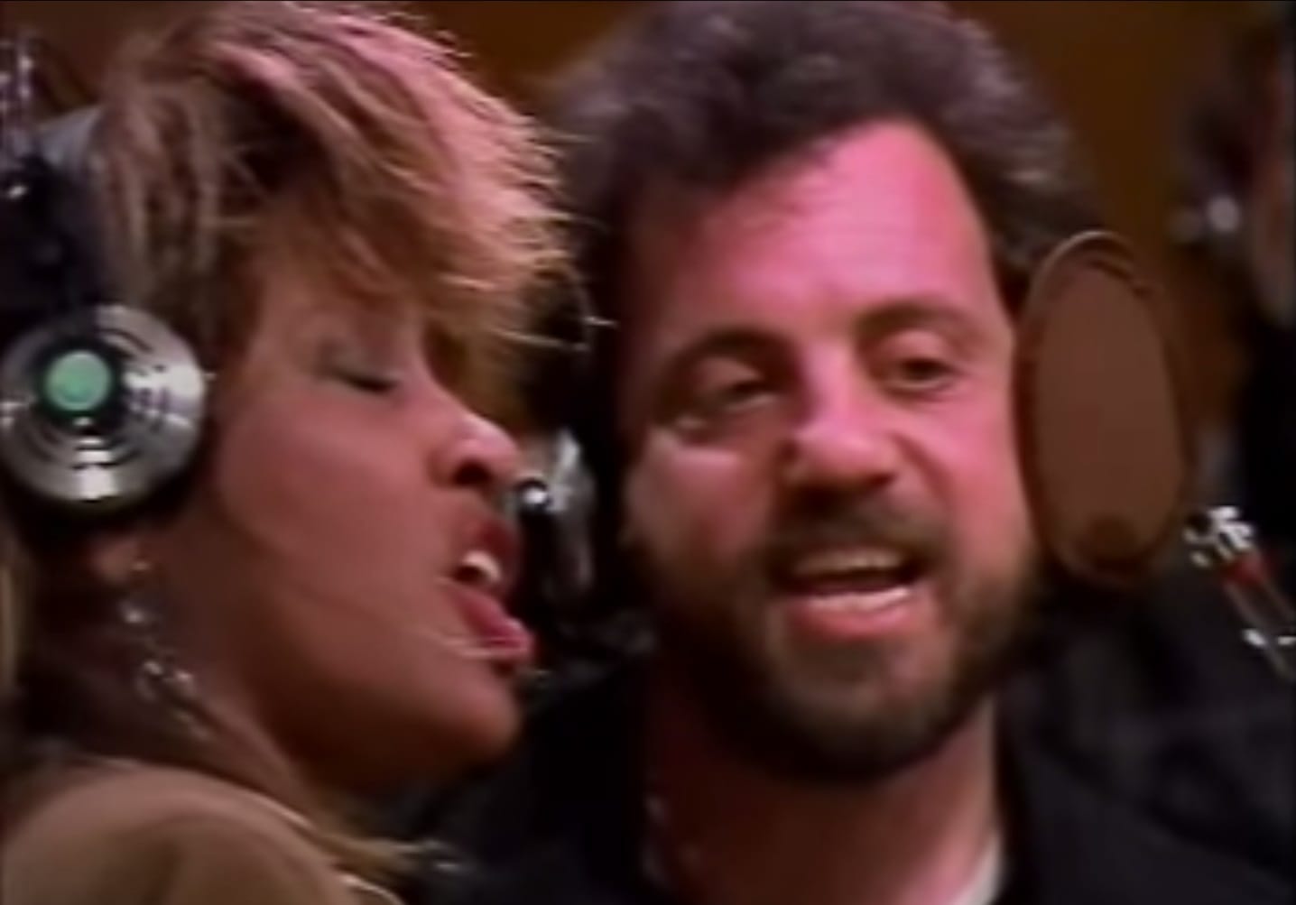 Tina Turner a Billy Joel se sluchátky na uších zpívají u mikrofonu při nahrávání We Are the World
