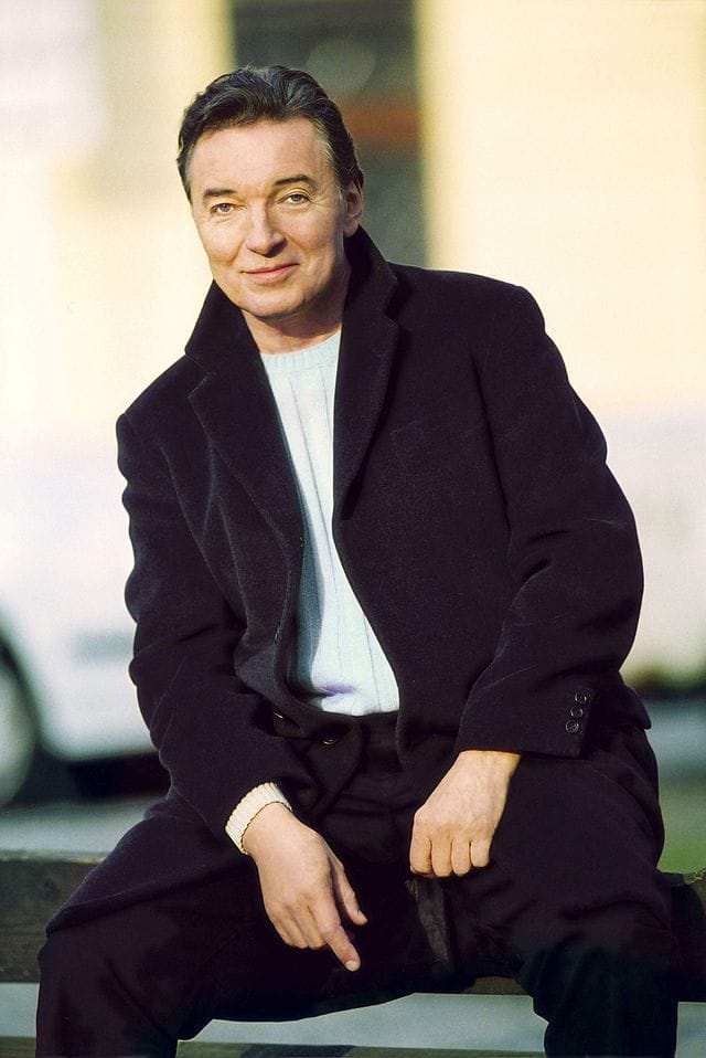 Karel Gott v černém kabátě a světlém svetru sedí na kraji lavičky a usmívá se