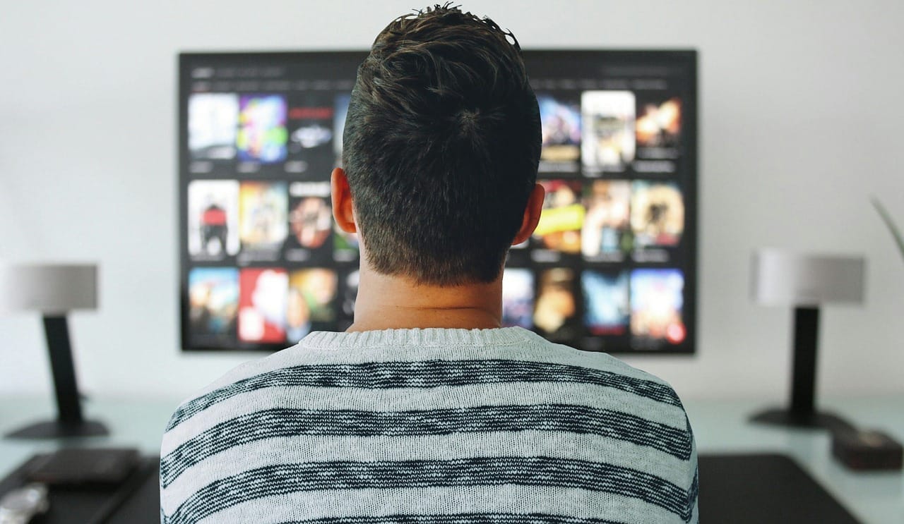Muž v pruhovaném svetru zezadu před televizí s nabídkou filmů a seriálů