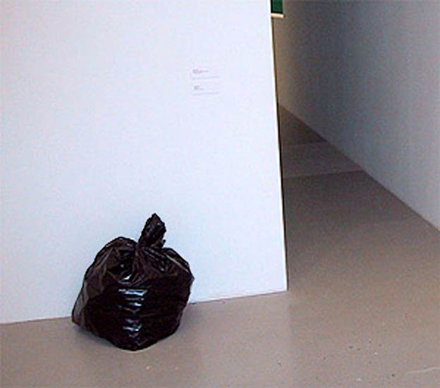 Zavázaný černý pytel na odpadky stojící u bílé stěny v prázdné galerijní chodbě