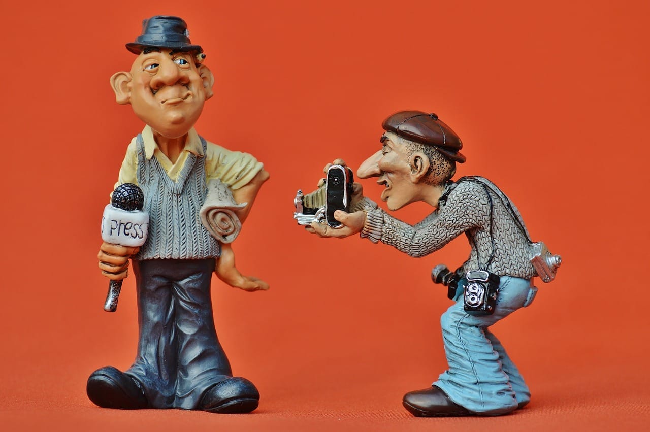 Dvě karikaturní figurky na oranžovém pozadí: novinář s mikrofonem s nápisem „press“ a fotograf fotící ho starým aparátem
