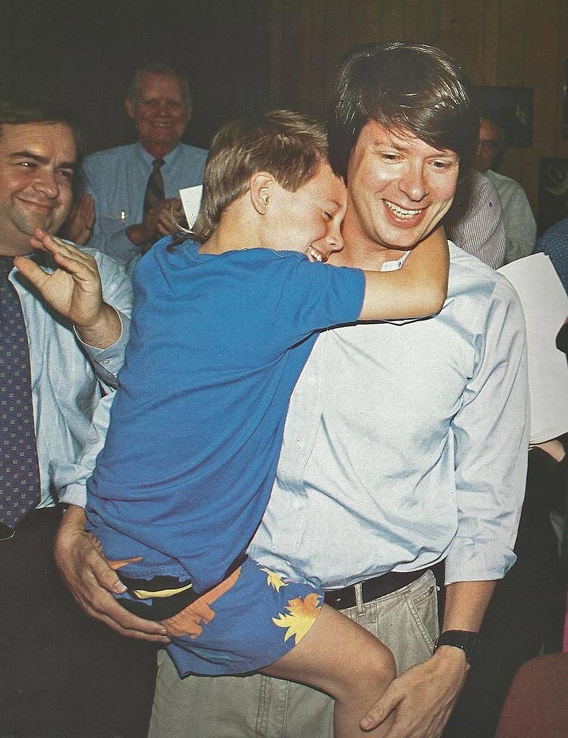 Usmívající se Dave Barry drží v náručí syna Roba v okamžiku, kdy se dozvěděl o Pulitzerově ceně, kolem tleskají lidé