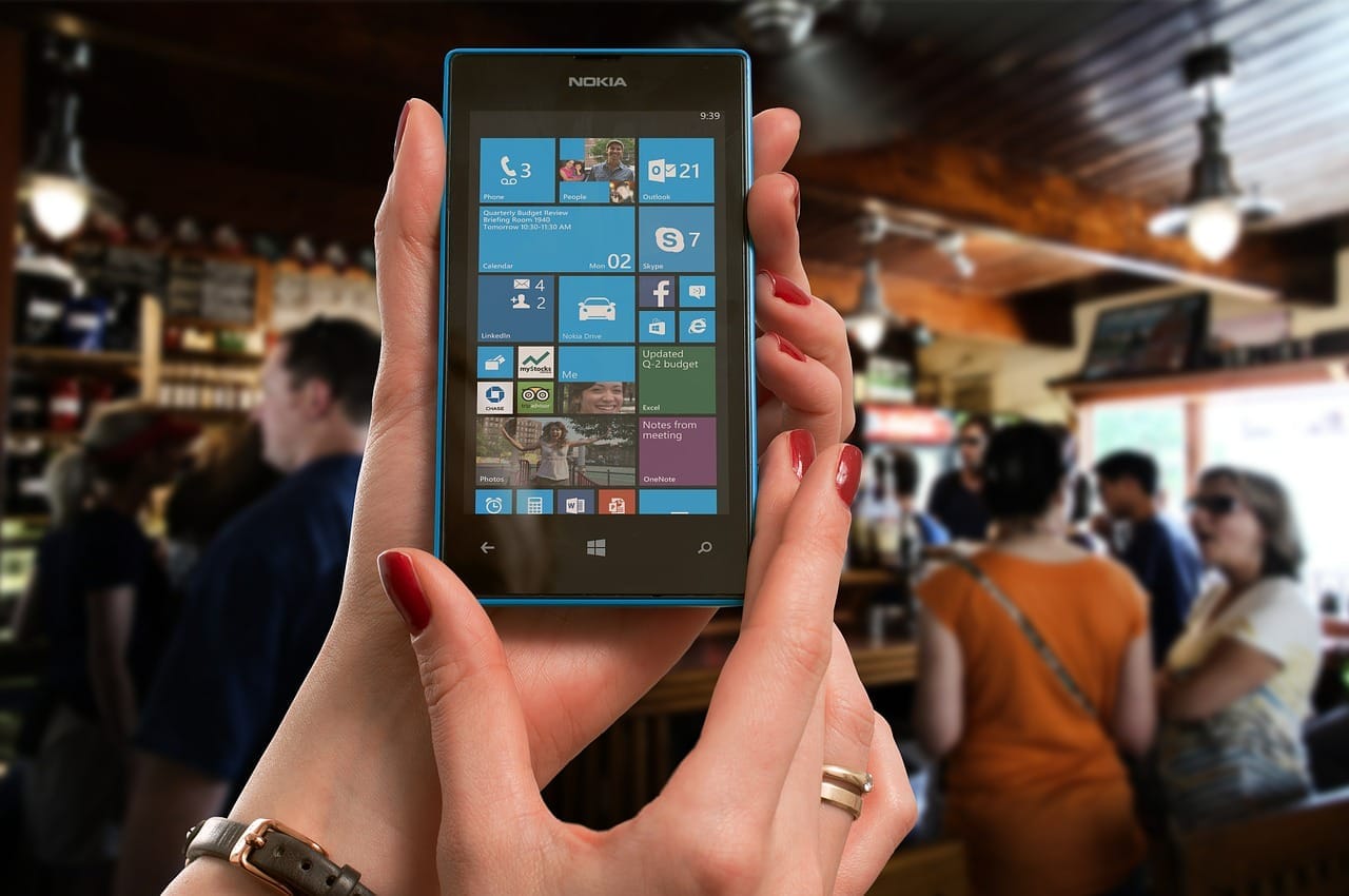 Ruce držící modrý telefon Nokia s dlaždicovou obrazovkou Windows Phone, v pozadí rozostřený interiér podniku s lidmi