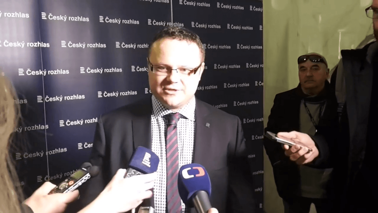 René Zavoral odpovídá novinářům před stěnou s logy Českého rozhlasu, u úst má mikrofony rozhlasu a České televize