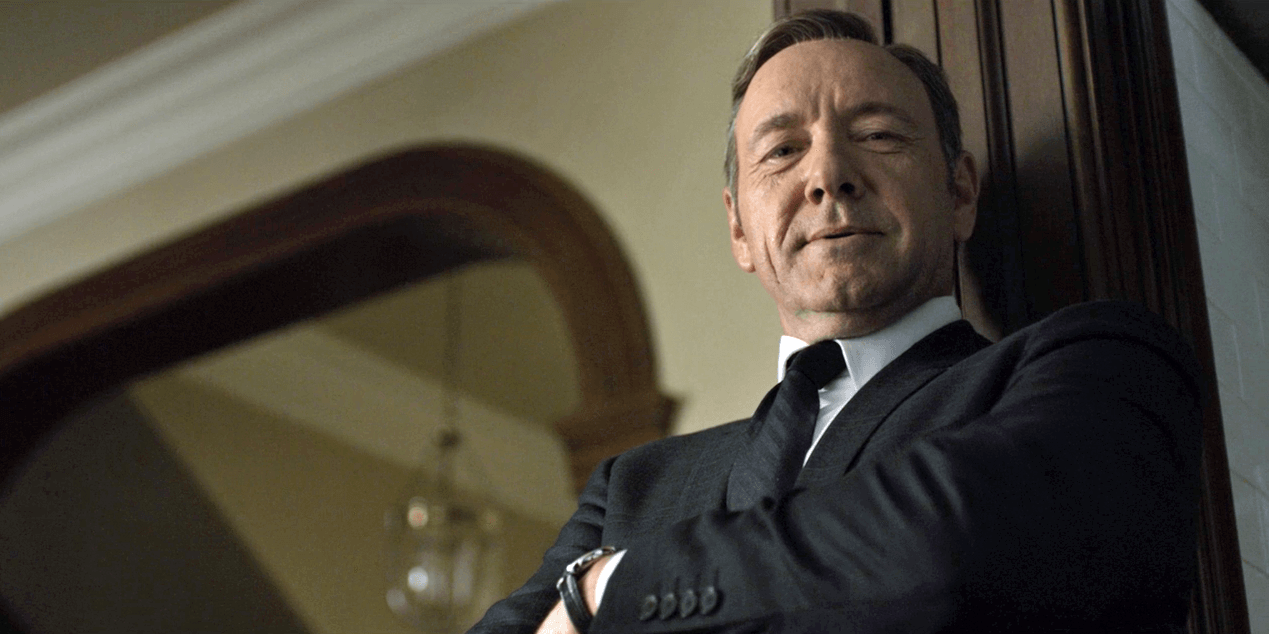 Kevin Spacey jako Frank Underwood v tmavém obleku s kravatou, opřený o zábradlí, hledí do kamery