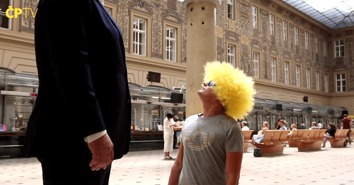 Záběr z marketingového videa České pošty: muž ve žluté kudrnaté paruce vzhlíží vzhůru v hale hlavní pošty, vlevo postava v obleku a v rohu logo ČPTV