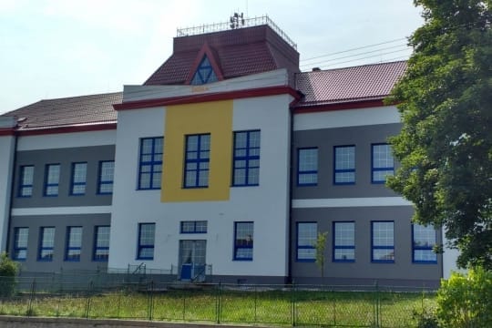 Zrekonstruovaná školní budova s bílo-šedou fasádou, žlutým pásem uprostřed a modrými okenními rámy za plotem