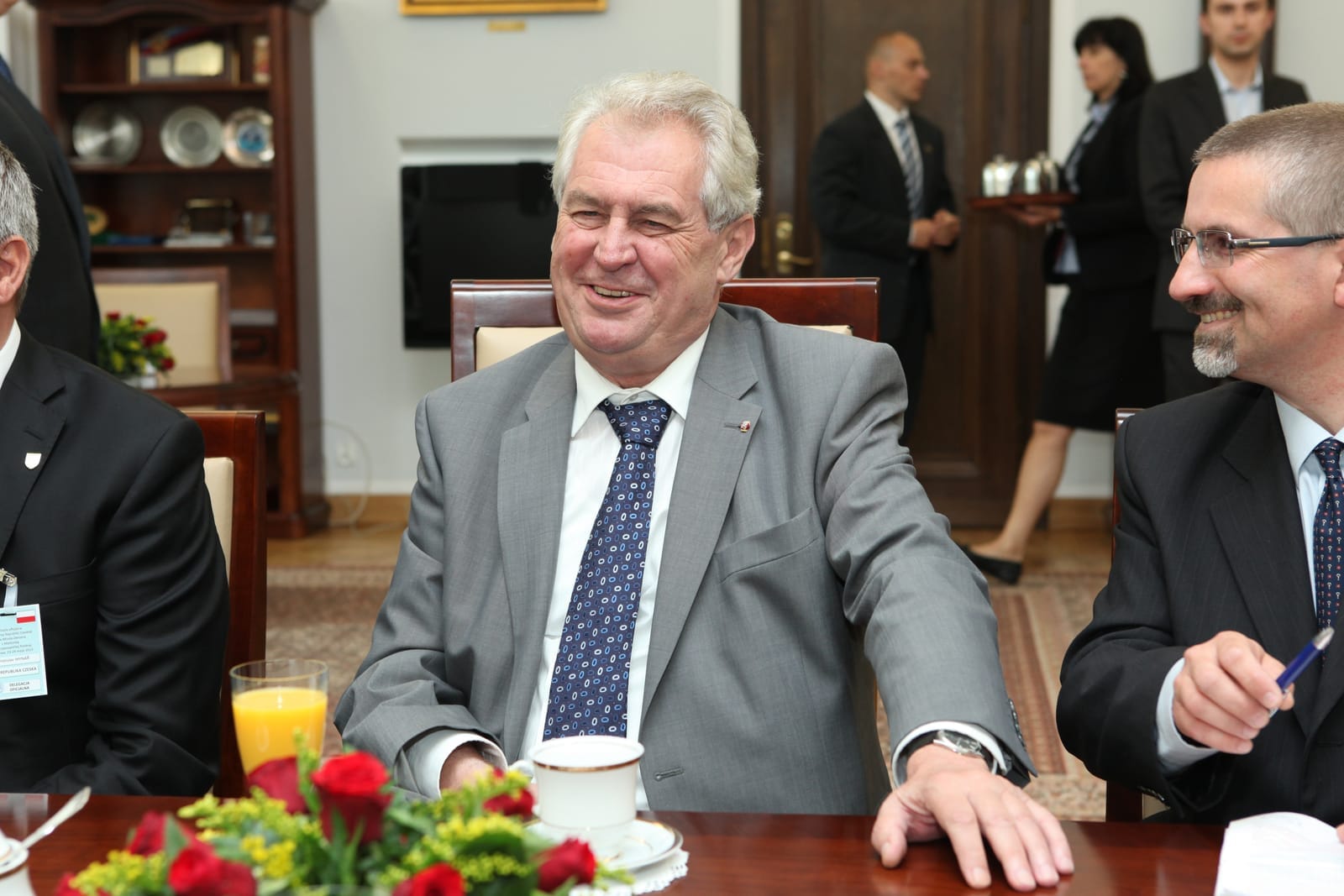 Miloš Zeman se smíchem sedí u stolu s růžemi, sklenicí pomerančového džusu a šálkem kávy, vedle něj usměvavý muž s brýlemi