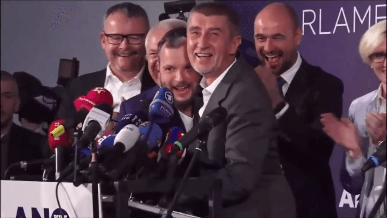 Andrej Babiš se smíchem objímá Marka Prchala u pultu s mikrofony, kolem stojí usmívající se lidé před logem ANO