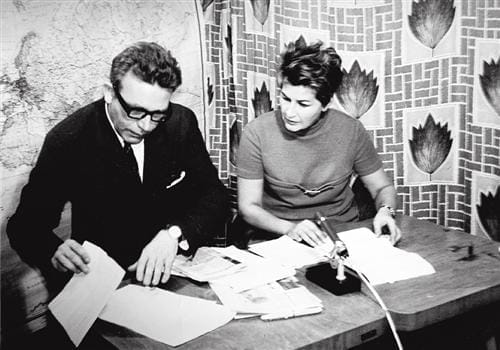 Kamila Moučková s krátkými vlasy a kolega v brýlích u stolu s papíry a mikrofonem během vysílání Československé televize v roce 1968, v pozadí mapa a vzorovaný závěs
