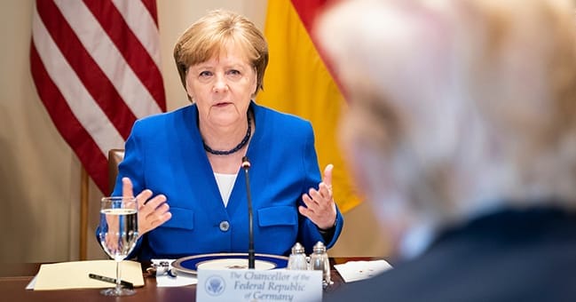 Angela Merkelová v modrém sáčku gestikuluje u jednacího stolu, před ní cedulka Chancellor of the Federal Republic of Germany