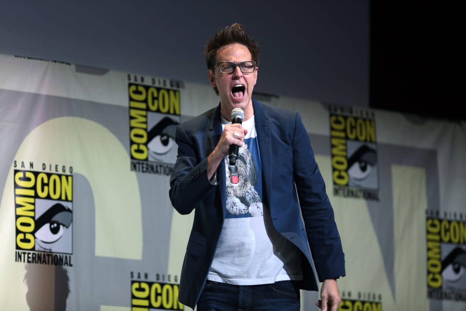 James Gunn s mikrofonem křičí na pódiu před stěnou s logy San Diego Comic-Con International