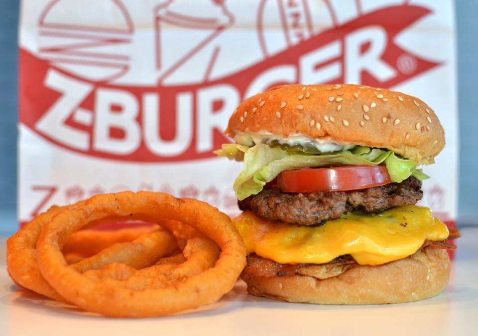 Cheeseburger se salátem a rajčetem, vedle smažené cibulové kroužky, v pozadí papírový sáček s logem Z-Burger