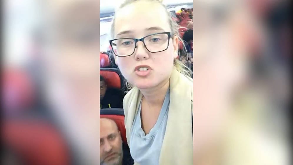 Mladá žena s brýlemi mluví do kamery mobilu v uličce letadla mezi červenými sedadly s cestujícími