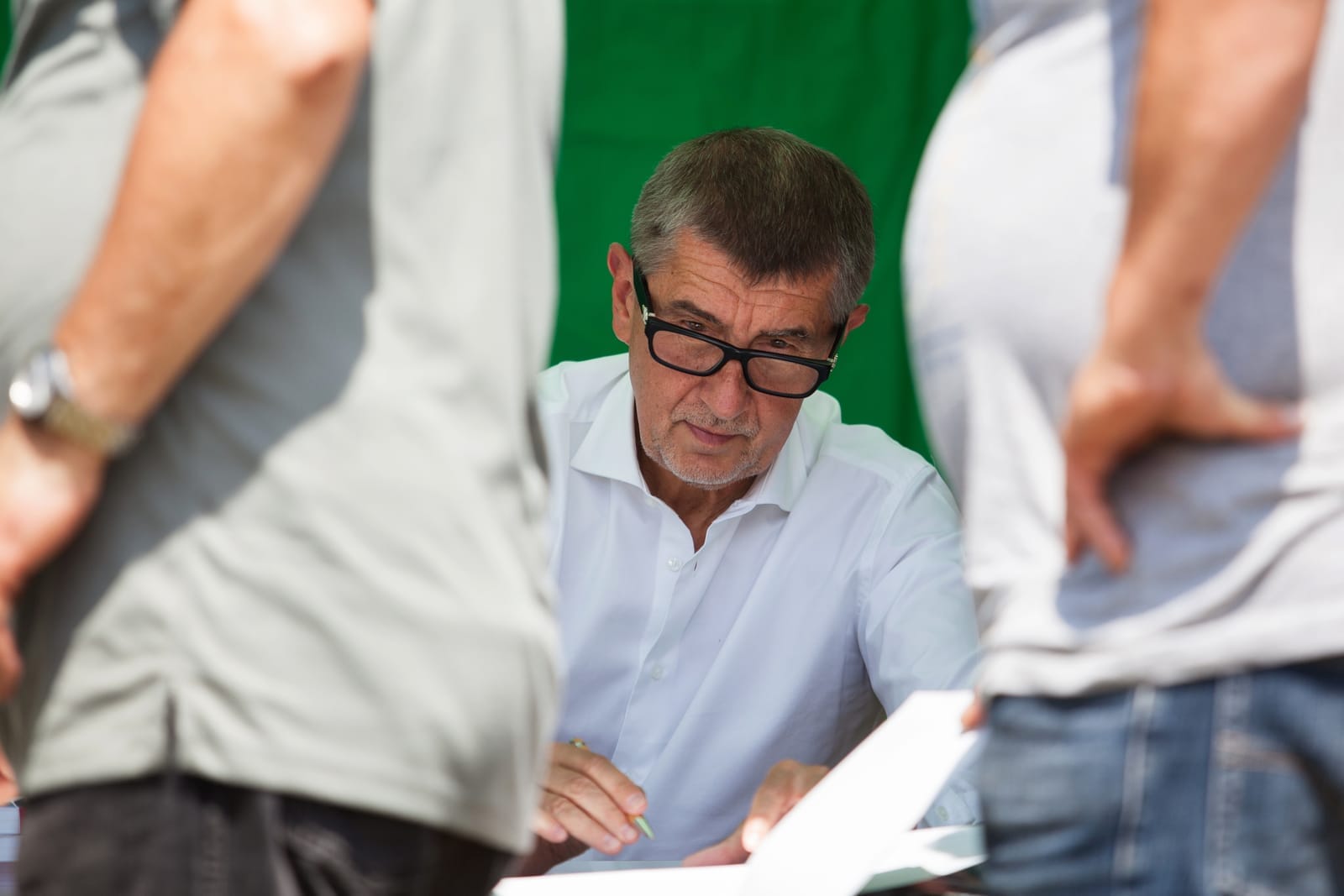 Andrej Babiš v brýlích a bílé košili podepisuje papír, v popředí rozmazané postavy stojících lidí