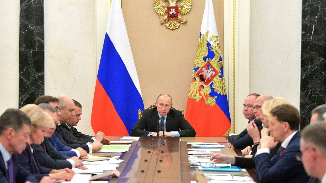 Vladimir Putin sedí v čele dlouhého jednacího stolu, za ním ruská vlajka, prezidentská standarta a státní orel