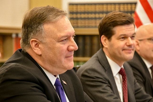 Mike Pompeo v obleku sedí u jednacího stolu vedle dalších dvou mužů a americké vlajky