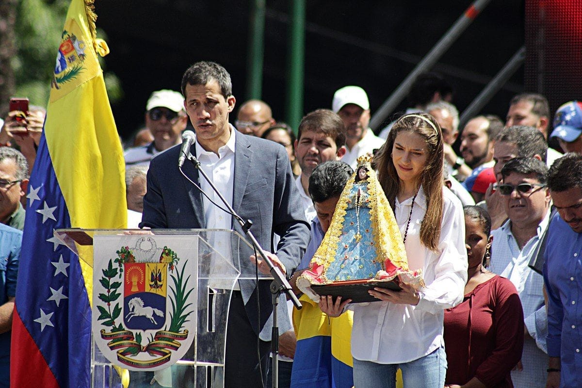 Juan Guaidó mluví do mikrofonu za skleněným pultem s venezuelským státním znakem, vedle žena držící sošku Madony