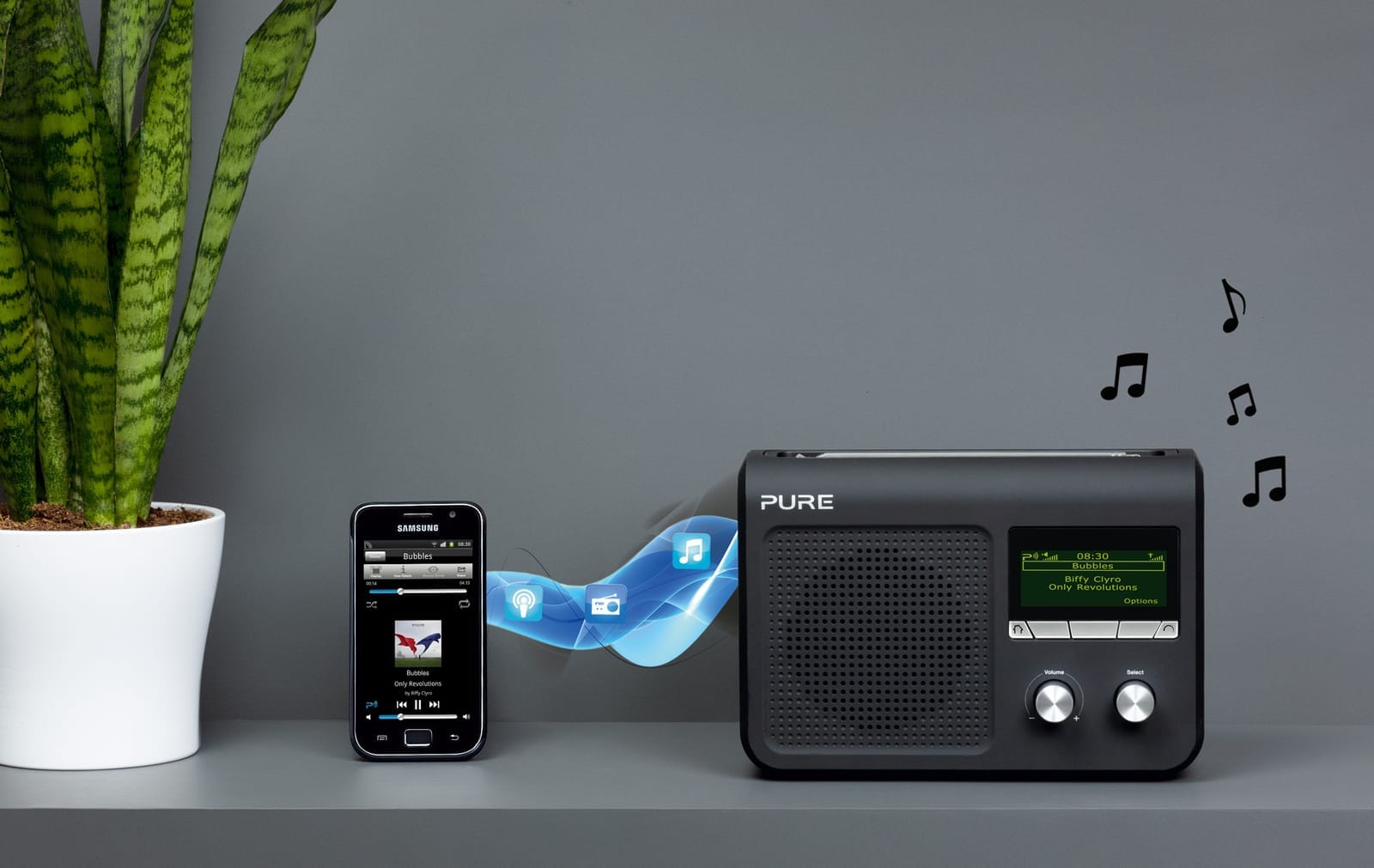 Černé digitální rádio Pure na poličce vedle mobilu Samsung s přehrávačem hudby, mezi nimi symbol bezdrátového přenosu