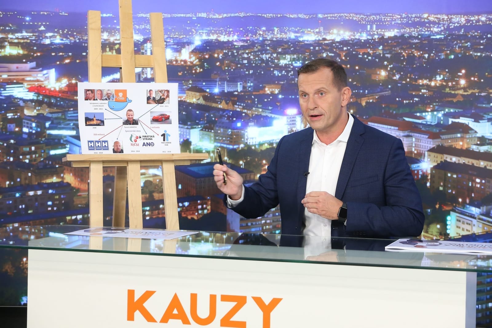Jaromír Soukup gestikuluje ve studiu s panoramatem noční Prahy, vedle stojanu se schématem vazeb a nápisem KAUZY na pultu