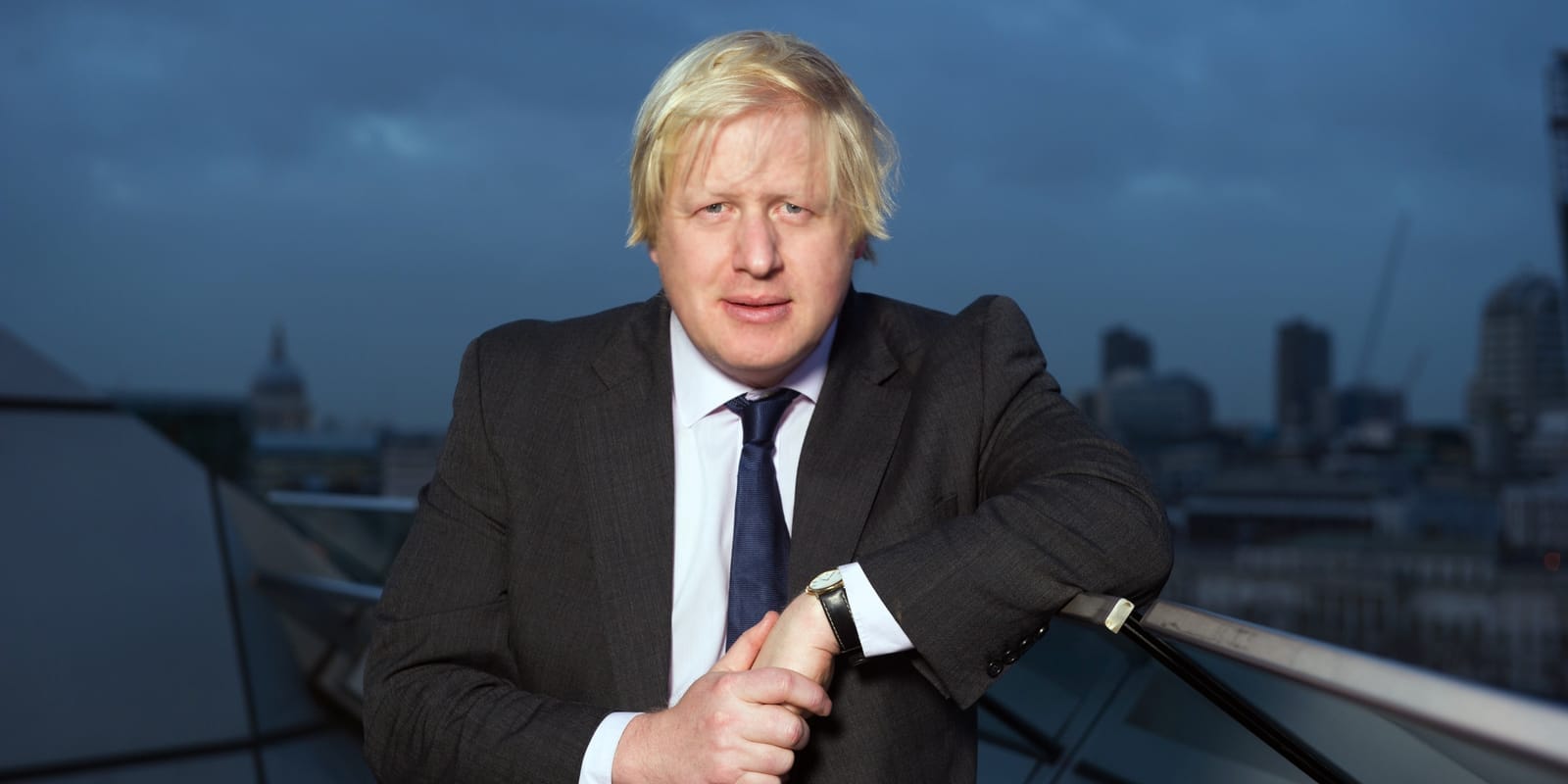 Boris Johnson v tmavém obleku s modrou kravatou se opírá o zábradlí nad panoramatem Londýna