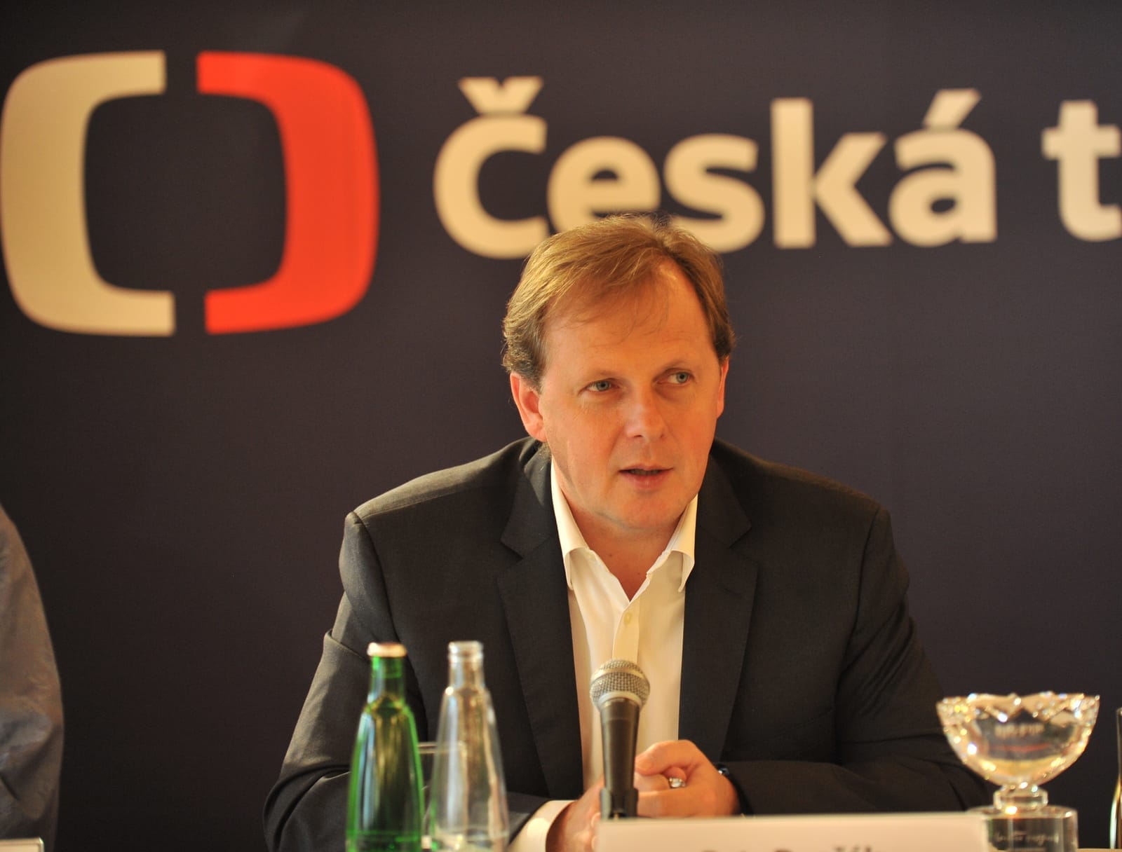 Petr Dvořák mluví do mikrofonu na tiskové konferenci před logem České televize