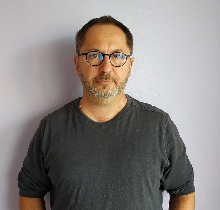 Petr Kukal s brýlemi, krátkými vlasy a prošedivělým strništěm v tmavošedém tričku před bílou zdí