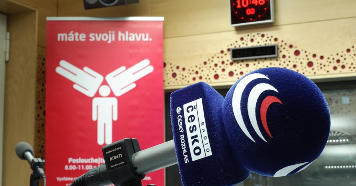 Modrý mikrofon s logem Radia Česko ve studiu, v pozadí červený banner s textem „máte svoji hlavu. Poslouchejte 8.00–11.00“ a digitální hodiny 10:46