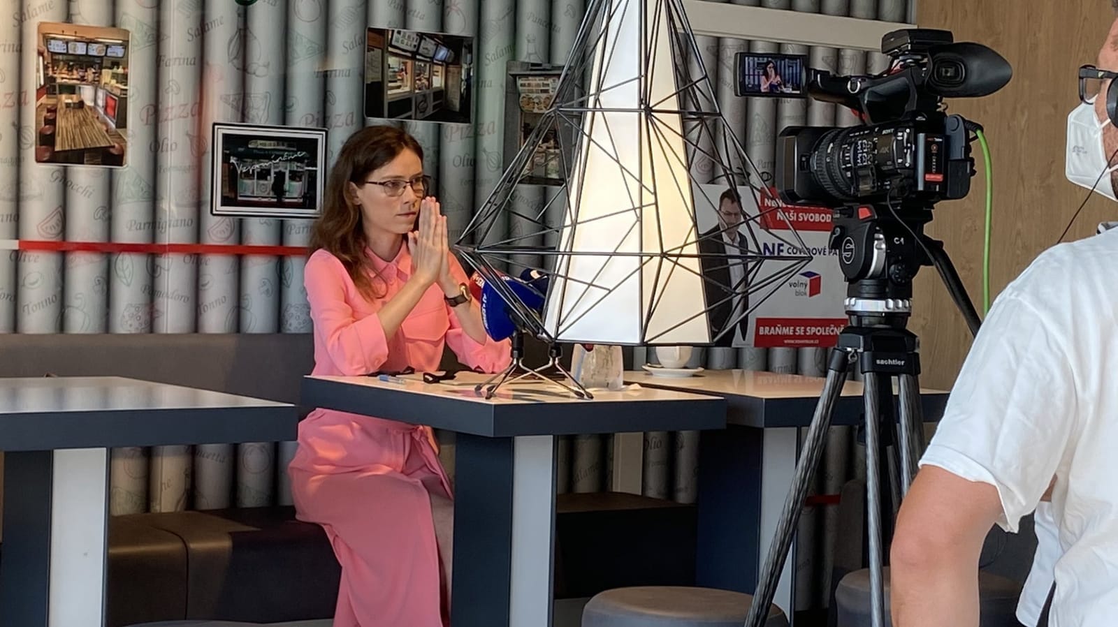 Hana Lipovská v růžové košili se sepjatýma rukama sedí u stolu v kavárně, vpravo kameraman u kamery na stativu