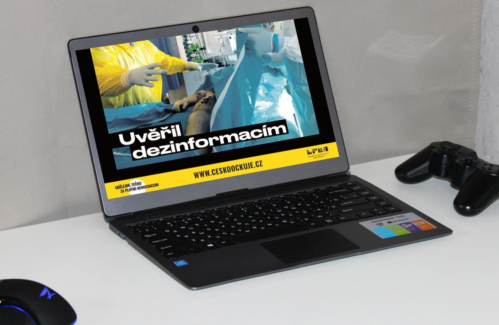 Notebook na stole zobrazuje kampaňový vizuál s lékaři u pacienta a nápisem „Uvěřil dezinformacím“ a adresou www.ceskoockuje.cz