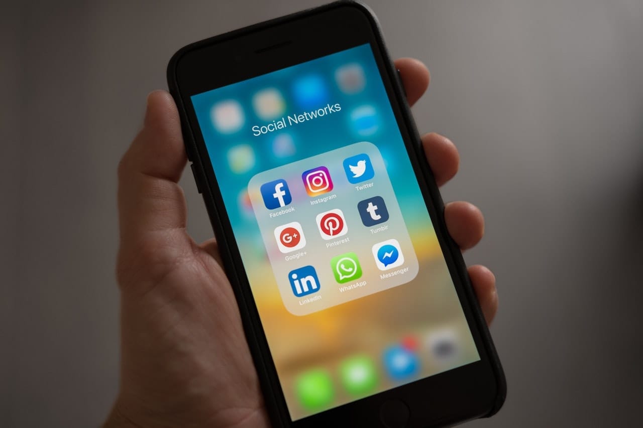 Ruka drží mobilní telefon s otevřenou složkou „Social Networks“ s ikonami Facebooku, Instagramu, Twitteru, Googlu+, Pinterestu, Tumblru, LinkedInu, WhatsAppu a Messengeru