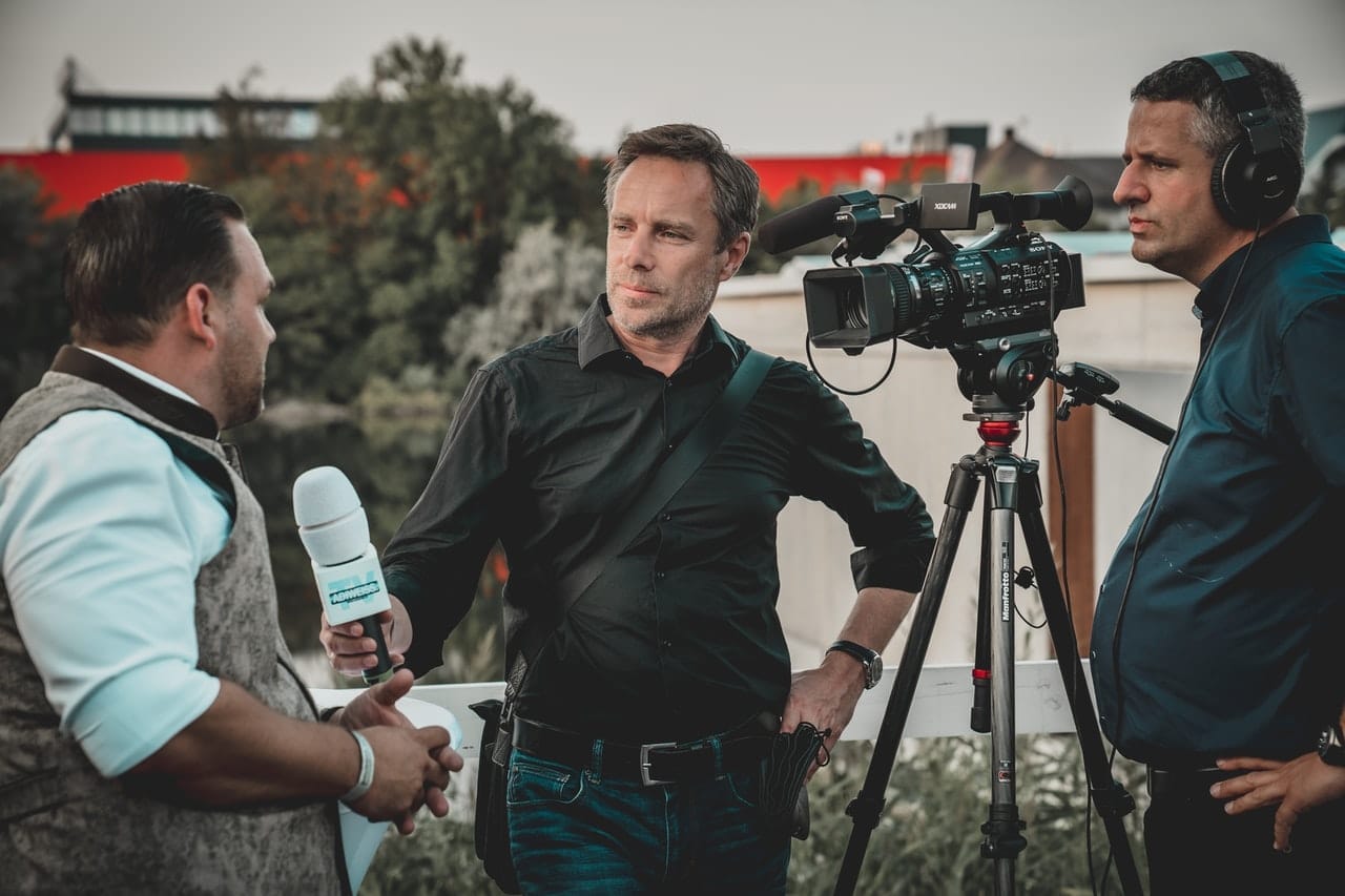 Reportér s mikrofonem hovoří s mužem v černé košili, vedle stojí kameraman se sluchátky u kamery na stativu