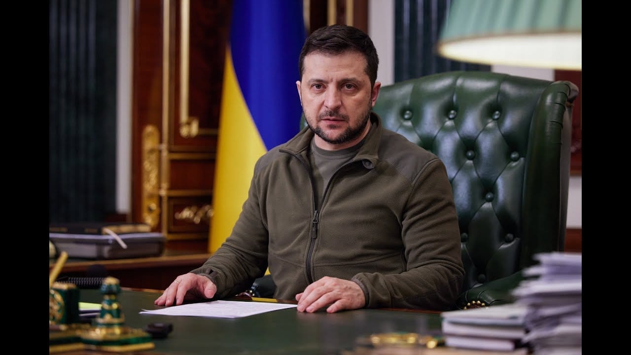 Volodymyr Zelenskyj v khaki fleecové bundě sedí za pracovním stolem před ukrajinskou vlajkou