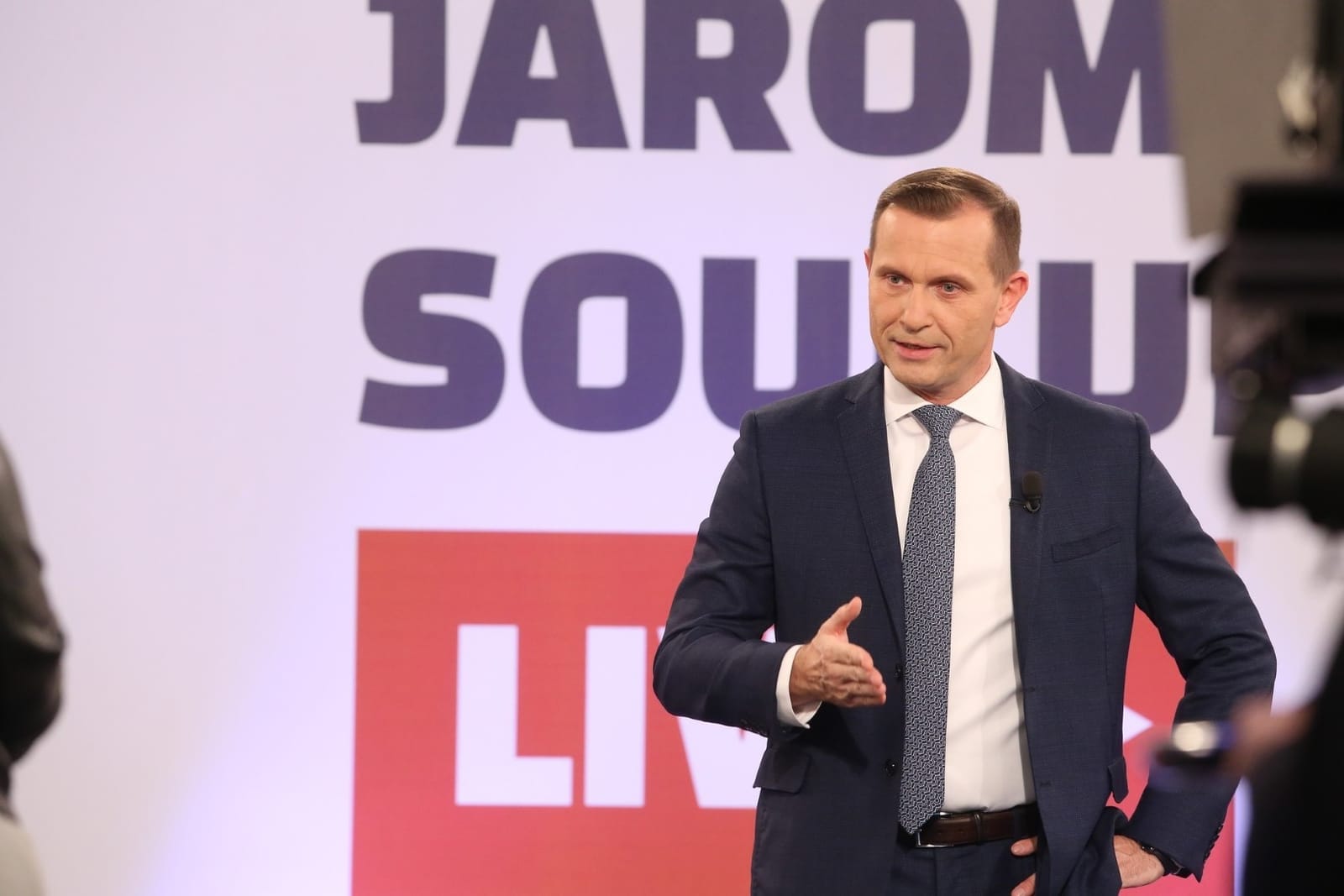 Jaromír Soukup v modrém obleku gestikuluje ve studiu před nápisem „Jaromír Soukup LIVE“