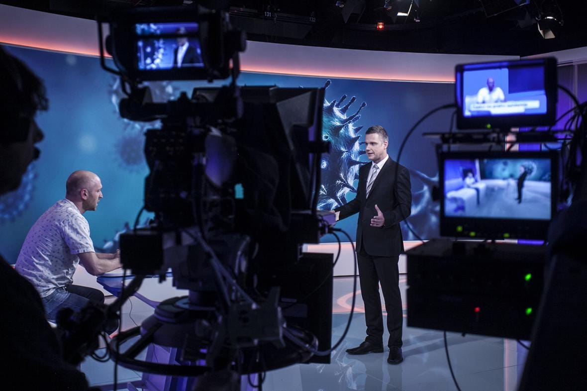 Daniel Stach v obleku moderuje ve studiu České televize s grafikou virů, v popředí kamera a monitory