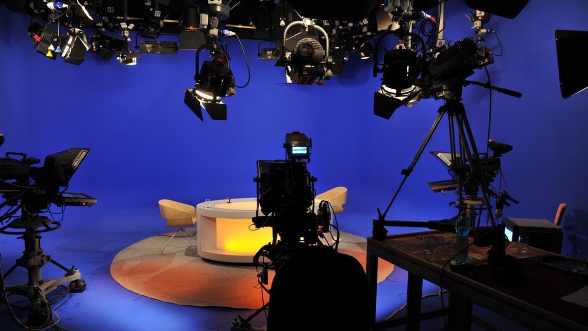Prázdné televizní studio s modrým pozadím, kamerami, reflektory a kruhovým stolem se dvěma křesly
