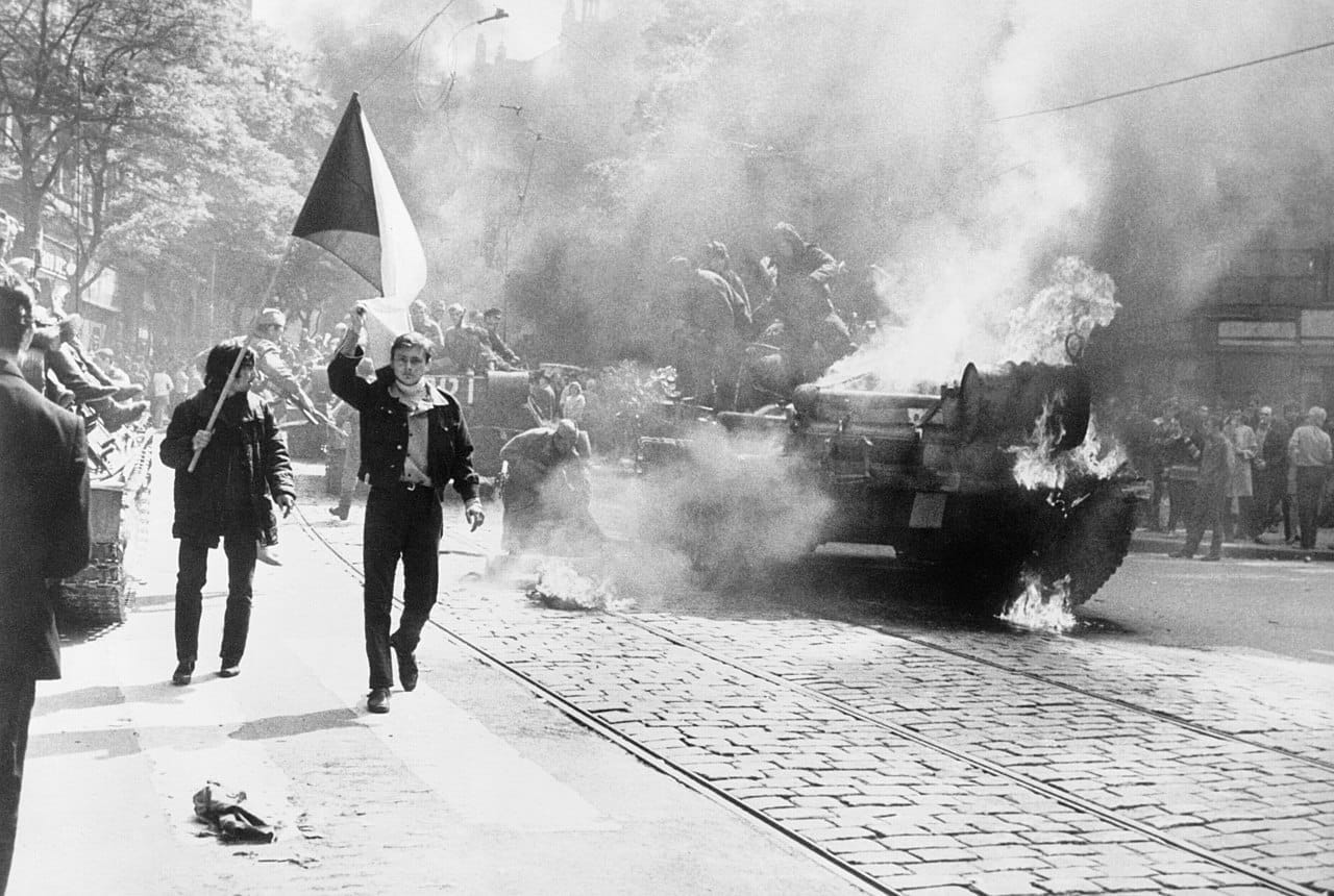 Černobílý snímek z pražské ulice v srpnu 1968: mladík nese československou vlajku, vedle na kolejích hoří tank obklopený kouřem a lidmi