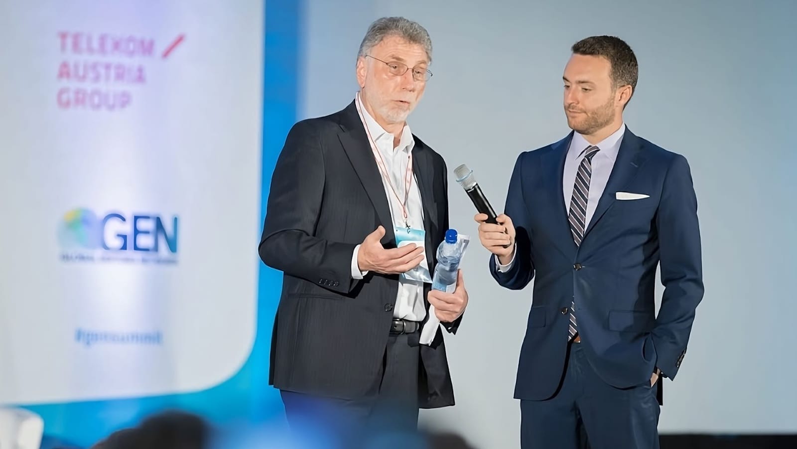 Martin Baron s brýlemi hovoří na pódiu, moderátor mu drží mikrofon před bannerem s logy Telekom Austria Group a GEN