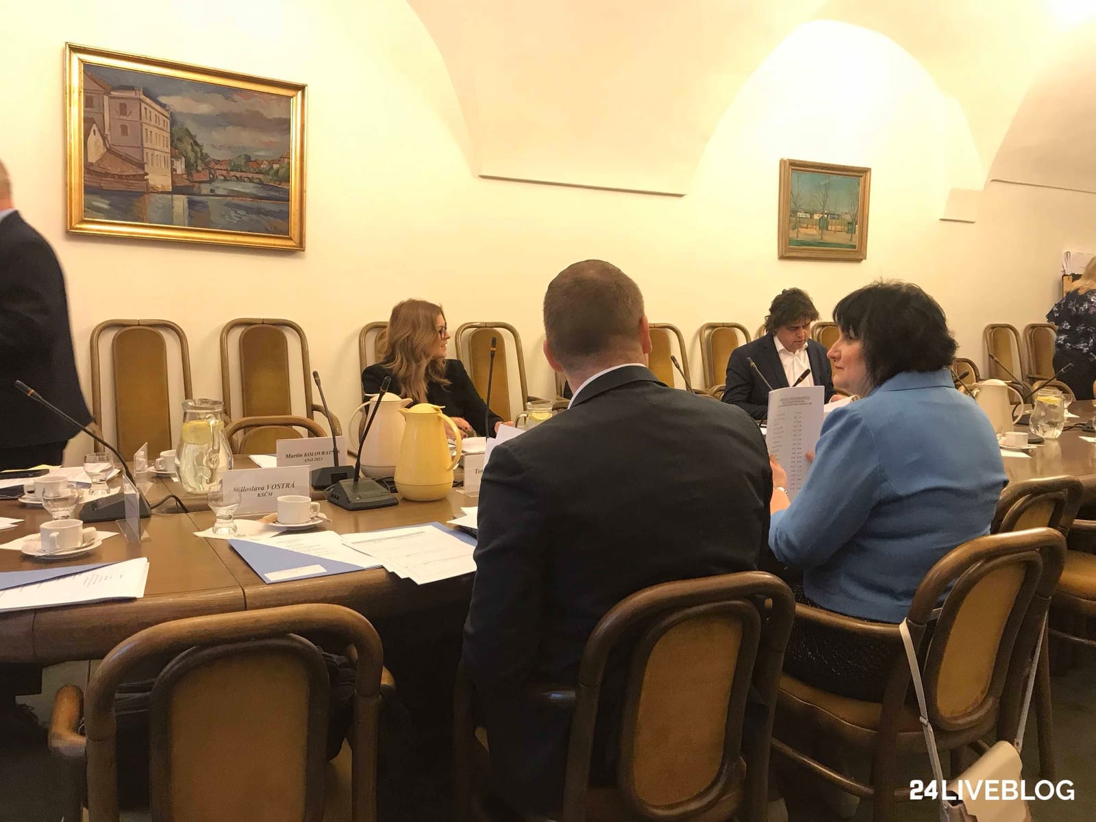 Zasedací místnost s klenutým stropem, kolem stolu s mikrofony, šálky a jmenovkami poslanců sedí několik lidí, na stěnách dvě zarámované krajinomalby; vpravo dole vodoznak 24LIVEBLOG