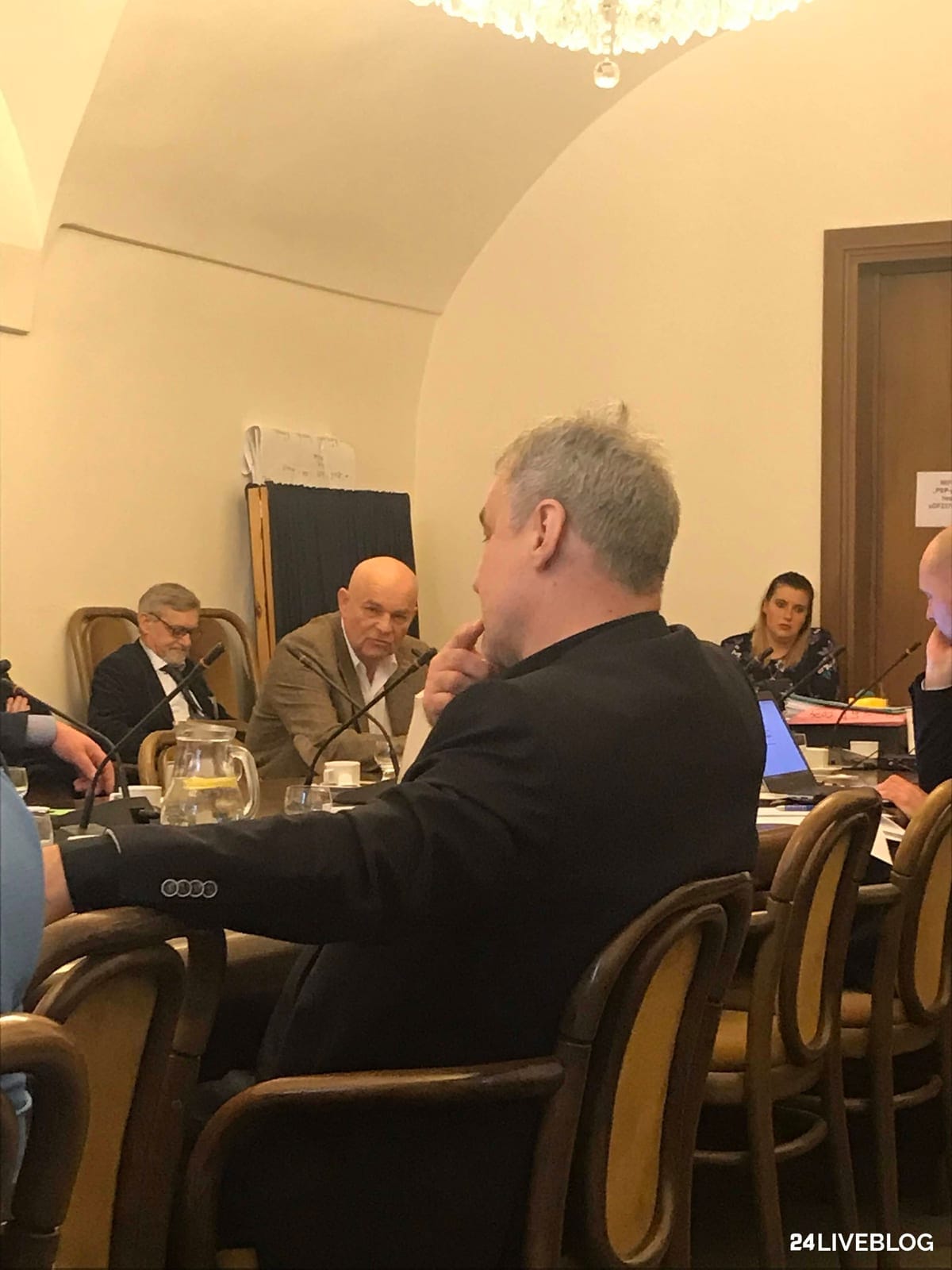 Muž v tmavém saku zády k objektivu mluví do mikrofonu, naproti němu u stolu sedí dva muži a poslouchají, vpravo v pozadí další účastníci jednání; vpravo dole vodoznak 24LIVEBLOG